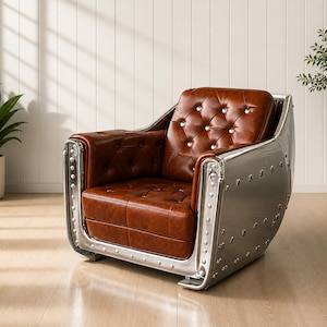 Op de afbeelding: Bruin lederen fauteuil met een gecapitonneerde rugleuning en zitting, geplaatst in een geklonken, zilverkleurig metalen frame. De stoel heeft een uniek ontwerp, met een gebogen rug en armleuningen, en staat op een houten vloer.