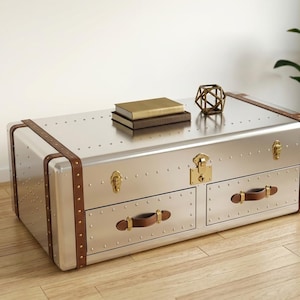Puede incluir: Una mesa de centro de metal plateado estilo ba&uacute;l con detalles en cuero marr&oacute;n y herrajes de lat&oacute;n. La mesa tiene dos cajones con tiradores de cuero y est&aacute; adornada con remaches. Dos libros y un objeto geom&eacute;trico dorado descansan encima.