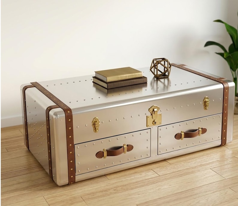 Puede incluir: Una mesa de centro de metal plateado estilo ba&uacute;l con detalles en cuero marr&oacute;n y herrajes de lat&oacute;n. La mesa tiene dos cajones con tiradores de cuero y est&aacute; adornada con remaches. Dos libros y un objeto geom&eacute;trico dorado descansan encima.
