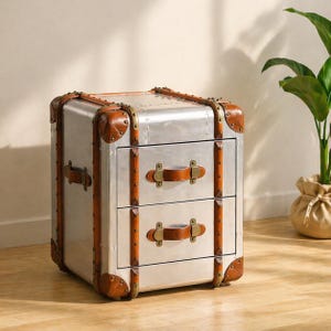 Op de afbeelding: Een zilverkleurige nachtkast met twee lades en bruin lederen accenten en handgrepen. De hoeken zijn versterkt met bruin leer en messingkleurige klinknagels. De nachtkast heeft een vintage design in de stijl van een reiskoffer.