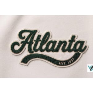 May include: A white fabric surface featuring a dark green chenille patch with the word Atlanta in a cursive script font. The patch includes a curved underline containing the text EST. 1847 in a smaller, clean, white serif typeface.