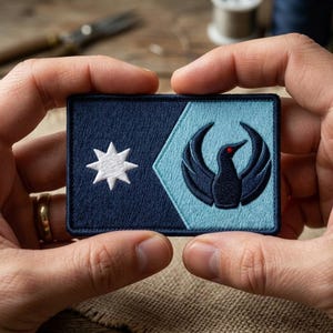 May include: A rectangular patch with a dark blue and light blue design. The left side features a white star, while the right side showcases a black bird emblem with a red eye. The patch is held between two hands.