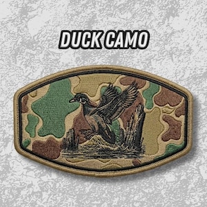 May include: Embroidered patch with a duck camo design. The patch features a duck in flight over water, with a camouflage background in shades of green, brown, and tan. The text "DUCK CAMO" is displayed in a bold, white font.