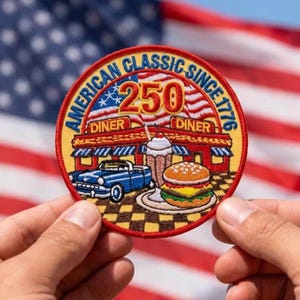 May include: A circular patch with the text "AMERICAN CLASSIC SINCE 1776" and "250 DINER". The patch features a diner, a blue convertible, a milkshake, and a burger. The background is the American flag.
