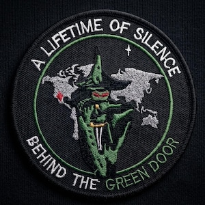 May include: Embroidered patch with the text "A LIFETIME OF SILENCE BEHIND THE GREEN DOOR." The patch features a green figure in a witch hat, a world map, and a dagger. The patch is circular with a black background.