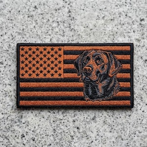 May include: A rectangular patch featuring an American flag design in shades of orange and black. The flag's stars are in the upper left corner, and a detailed dog portrait is on the right side. The patch is likely made of fabric.