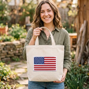 May include: A beige canvas tote bag featuring a rectangular embroidered patch of the United States flag on the front. The flag design displays the traditional red, white, and blue colors with a blue field of white stars and horizontal red and white stripes.