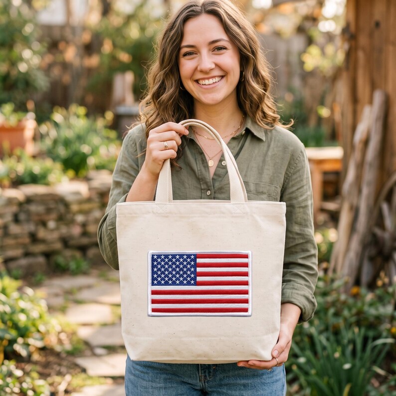 May include: A beige canvas tote bag featuring a rectangular embroidered patch of the United States flag on the front. The flag design displays the traditional red, white, and blue colors with a blue field of white stars and horizontal red and white stripes.