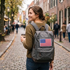May include: A gray canvas backpack featuring a rectangular patch of the United States flag on the front pocket. The backpack has a top handle, adjustable shoulder straps, and dark brown leather-like accents on the zippers and base.