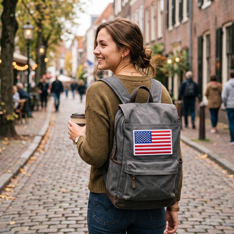 May include: A gray canvas backpack featuring a rectangular patch of the United States flag on the front pocket. The backpack has a top handle, adjustable shoulder straps, and dark brown leather-like accents on the zippers and base.