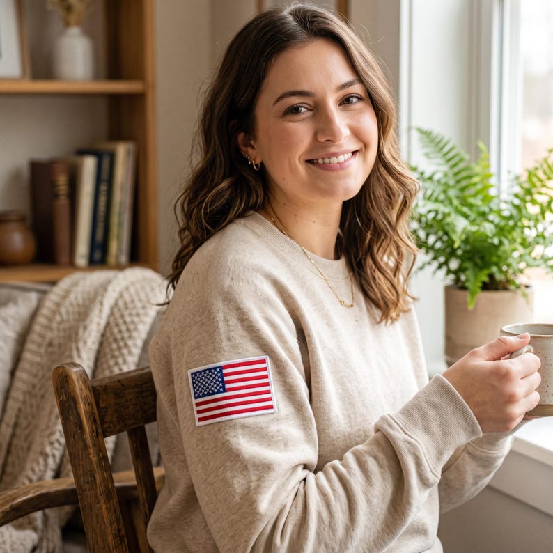 May include: A beige long-sleeve crewneck sweatshirt featuring a rectangular patch of the United States flag on the upper right sleeve. The garment is made of a soft, heathered fabric with ribbed cuffs and a ribbed neckline, providing a clean look.