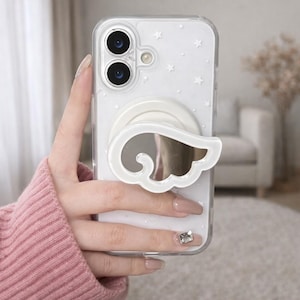 May include: Clear phone case with a white angel wing design and a mirrored surface. The case has a white pop-up grip and is decorated with small white stars. The phone case is held in a person's hand.