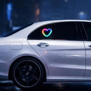 May include: A white car with a black wheel and a colorful heart-shaped light on the rear window. The heart has a gradient of colors, including yellow, orange, green, and blue. The car is parked at night.