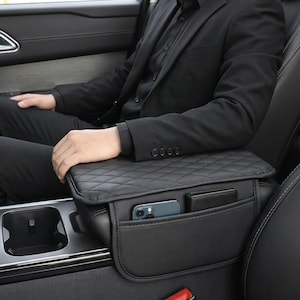 May include: Black leather car console organizer with a quilted top and multiple pockets. The organizer has a pocket holding a smartphone and another holding a wallet. The console is installed in a car.