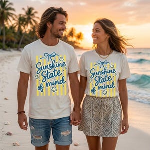 K&ouml;nnte beinhalten: Zwei Personen tragen passende cremefarbene T-Shirts mit dem Text "Sunshine State of Mind" in blauer Schrift und gelben Akzenten. Die Shirts haben eine Schleife und eine Blumen-Grafik. Sie halten sich am Strand an den H&auml;nden.