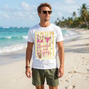 K&ouml;nnte beinhalten: Wei&szlig;es T-Shirt mit Strandmotiv. Das Design zeigt einen rosafarbenen Bikini auf einer W&auml;scheleine, eine Sonne, Sterne und den Schriftzug "Tanned and Tipsy". Das Shirt wird mit gr&uuml;nen Shorts an einem Sandstrand getragen.