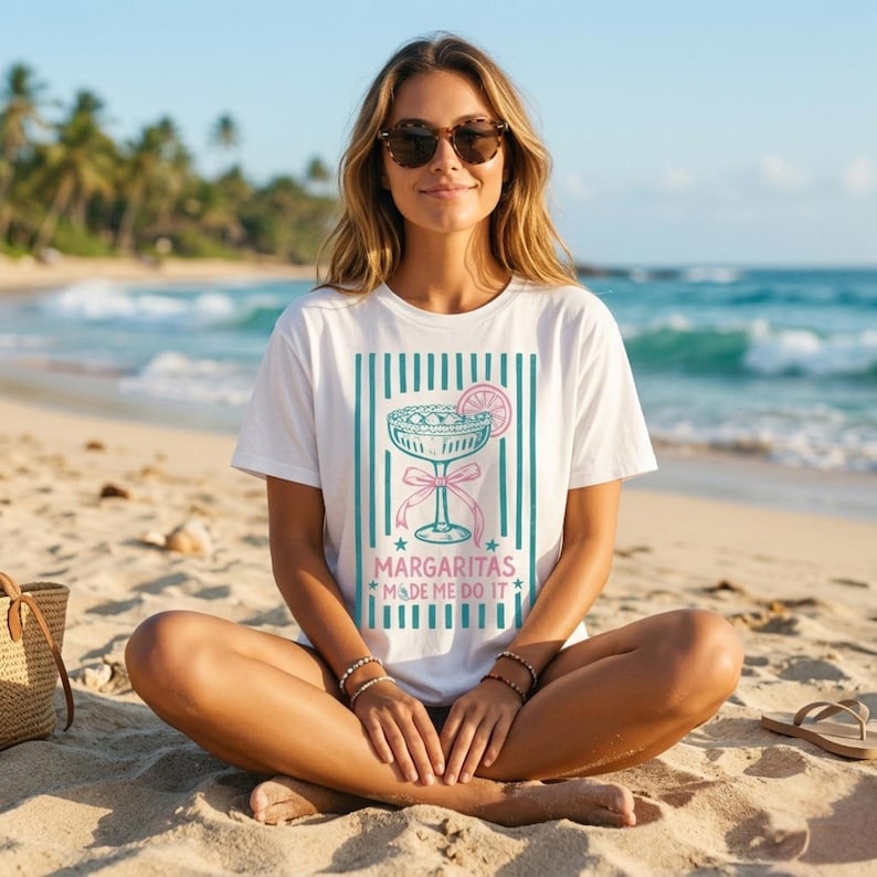 K&ouml;nnte beinhalten: Wei&szlig;es T-Shirt mit einer Grafik eines Margarita-Cocktails, einer rosa Schleife und dem Text "Margaritas Made Me Do It". Das Design befindet sich vor einem Hintergrund aus t&uuml;rkisfarbenen vertikalen Streifen. Das T-Shirt wird am Strand getragen.