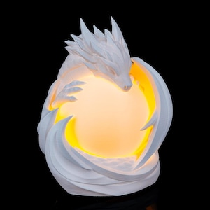 May include: A white dragon sculpture cradling a glowing orb. The dragon's detailed features include sharp spines and outstretched wings, with the orb emitting a warm, yellow light. The sculpture is set against a black background.