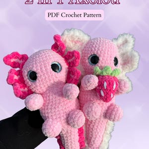 Könnte beinhalten: Zwei handgefertigte, rosa gehäkelte Axolotl, eines hält eine Erdbeere. Die Axolotl haben große, blau umrandete Augen und rosa Kiemen. Der Text "2 in 1 Axolotl" und "PDF Crochet Pattern" befindet sich oben im Bild.