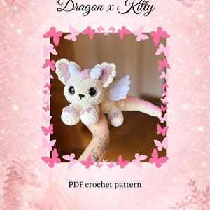 Könnte beinhalten: Ein gehäkeltes Drachen-Kitty-Hybrid in Weiß, Rosa und Hellblau, mit großen schwarzen Augen und kleinen Flügeln. Das Bild hat den Text "Dragon x Kitty" und "PDF crochet pattern". Der Hintergrund ist rosa mit Schmetterlings- und Blumenakzenten.