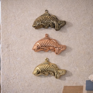 May include: Three decorative metal fish in bronze, copper, and gold tones are mounted on a textured wall. Each fish has detailed scales and fins, with a loop for hanging. The fish are arranged vertically, showcasing their different metallic finishes.