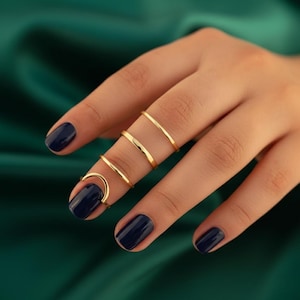 Puede incluir: Una mano con esmalte de uñas azul oscuro muestra una colección de anillos dorados. Los anillos incluyen una banda delgada, un anillo curvo y otras dos bandas simples. Los anillos se muestran sobre un fondo verde.