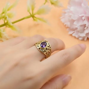 May include: Gold-colored ring with an oval, purple gemstone. The ring has an open filigree design around the gemstone and band. The ring is on a finger, with a soft-focus background of flowers.