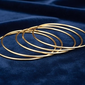 Puede incluir: Cinco pulseras finas de color dorado están dispuestas sobre una superficie de terciopelo azul oscuro. Las pulseras son circulares y parecen ser de metal. La joyería es sencilla y elegante, adecuada para el uso diario.