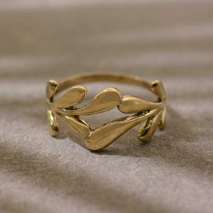 Peut inclure: Une bague dorée ornée d'un motif de feuilles. La bague est composée d'un anneau et est ornée de feuilles stylisées qui épousent le doigt. La bague est posée sur un fond neutre.