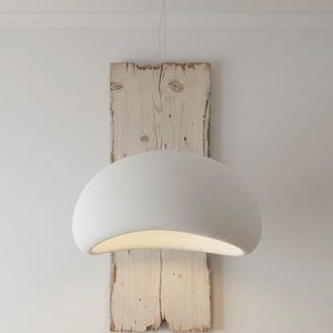 May include: A white, dome-shaped pendant light fixture hangs from a thin wire against a backdrop of weathered, light-colored wood planks. The light emits a soft glow, creating a warm ambiance. The fixture is mounted on a white wall.