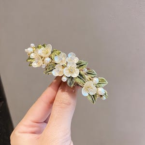 Könnte beinhalten: Ein goldfarbener Haarklammer mit weißen Blüten, grünen Blättern und Kunstperlen. Das florale Design ist mit kleinen Kristallen detailliert. Ein dekoratives Haaraccessoire.