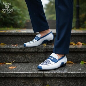 May include: White and blue leather loafers with a decorative floral design. The shoes have a classic slip-on style with a contrasting blue trim and sole. The image shows the shoes on a set of outdoor stairs.