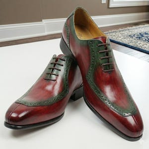 May include: A pair of burgundy leather dress shoes with dark green detailing. The shoes have an Oxford design with a cap toe and decorative perforations. Brown laces and black soles complete the look.