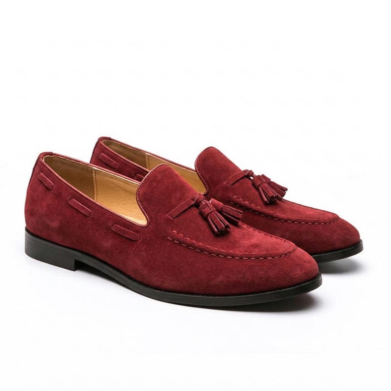 May include: A pair of burgundy suede loafers with tassel details. The shoes have a rounded toe and a black sole. The loafers are a slip-on style and are displayed on a white background.