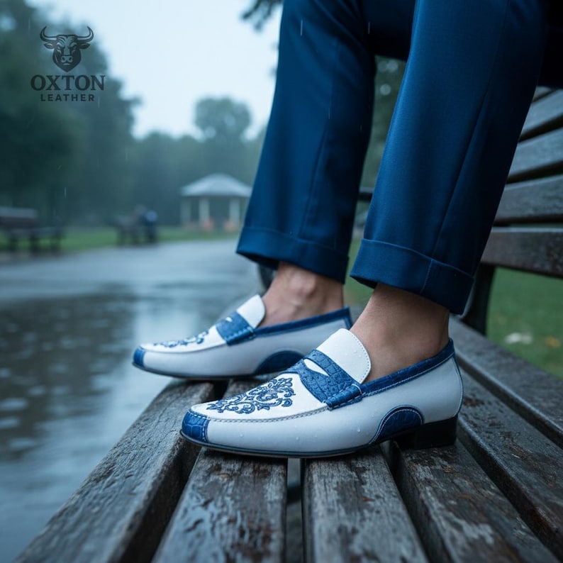 May include: White and blue leather loafers with intricate blue embroidery. The shoes have a classic design with a rounded toe and a slip-on style. The Oxton Leather logo is visible in the upper left corner.