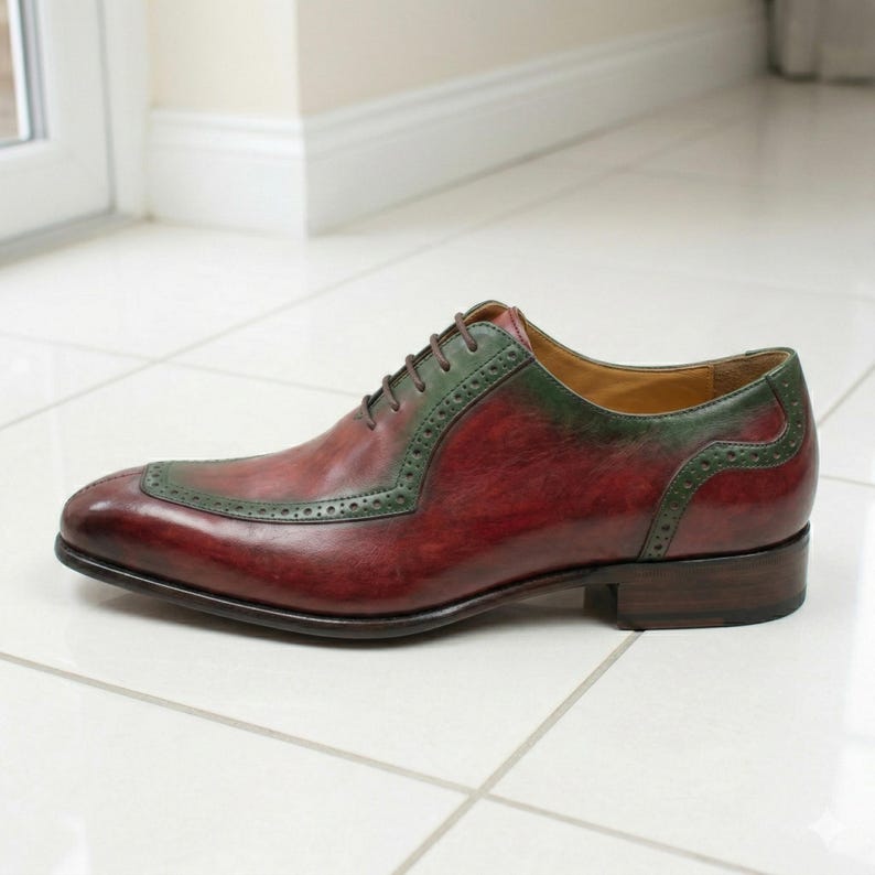 May include: A burgundy leather dress shoe with green detailing. The shoe has a wingtip design with decorative perforations. The laces and the top of the shoe are green, and the sole is brown.