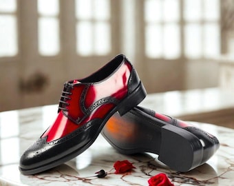 May include: A pair of two-tone dress shoes with a glossy red upper and black accents. The shoes feature a classic wingtip design with intricate detailing and a lace-up closure. The shoes are displayed on a marble surface.