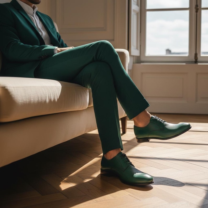 May include: A green suit with matching green leather dress shoes. The suit jacket is buttoned, and the person is seated on a beige sofa. The shoes have a classic lace-up design.