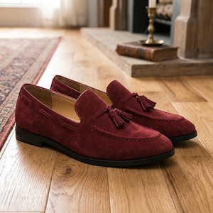 May include: A pair of burgundy suede loafers with tassel details. The shoes have a classic design with a rounded toe and a low heel. They are displayed on a wooden floor, suggesting a focus on footwear and fashion.
