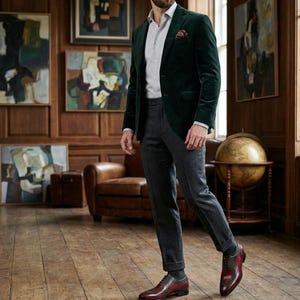 May include: A man's outfit featuring a dark green velvet blazer, grey trousers, and burgundy leather shoes. The ensemble includes a light grey shirt and a patterned pocket square. The room has wood paneling and artwork.