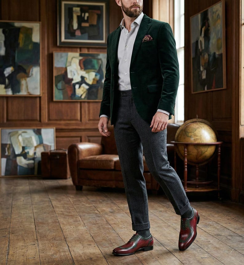 May include: A man's outfit featuring a dark green velvet blazer, grey trousers, and burgundy leather shoes. The ensemble includes a light grey shirt and a patterned pocket square. The room has wood paneling and artwork.