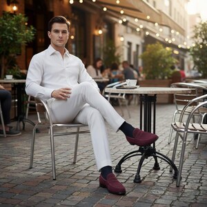 May include: A person in a white shirt and trousers sits at an outdoor cafe, wearing burgundy loafers and dark socks. The cafe has small round tables and metal chairs. The setting is a European street.