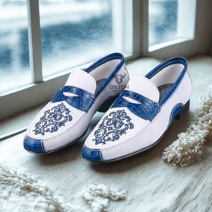May include: A pair of white leather loafers with blue accents. The shoes feature a blue crocodile-textured vamp and trim, with a decorative floral design on the toe. The brand name "OXTON LEATHER" is visible on the side.