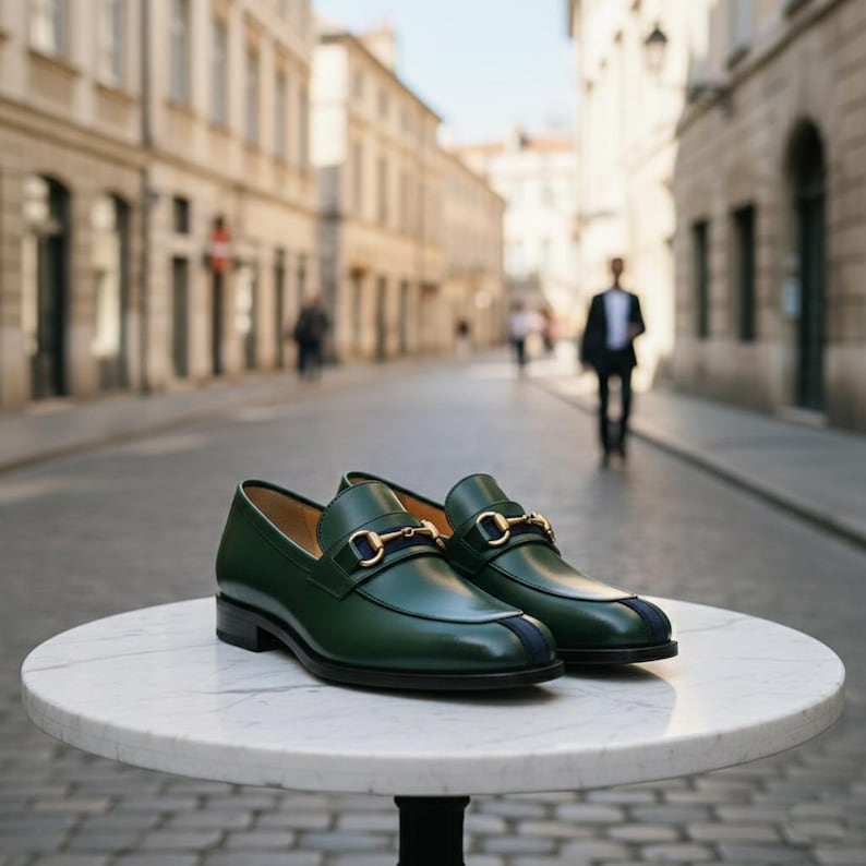 May include: A pair of green leather loafers with gold-colored metal accents and a navy blue stripe detail. The shoes are displayed on a white marble table, with a blurred city street in the background.