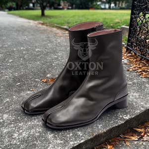May include: A pair of dark gray leather ankle boots with a split-toe design. The boots have a low heel and the text "OXTON LEATHER" is visible on the side. The boots are displayed on a concrete surface.