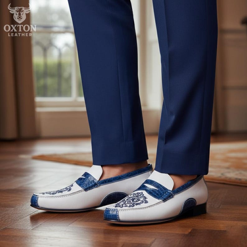 May include: White leather loafers with blue crocodile-embossed leather accents. The shoes feature intricate embroidered designs on the vamp and a contrasting blue trim. The shoes are paired with navy blue trousers.