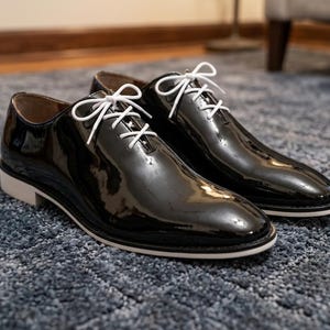Può includere: Un paio di scarpe eleganti in vernice nera con lacci e suole bianche. Le scarpe sono molto lucide, riflettendo la luce, e sono appoggiate su un tappeto blu. Il design classico suggerisce un abbigliamento formale.
