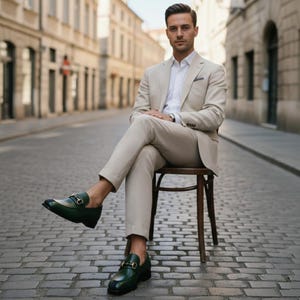 May include: A man in a beige suit and white shirt sits on a wooden chair on a cobblestone street. He wears dark green leather loafers with gold-colored accents. The suit jacket has a pocket square.