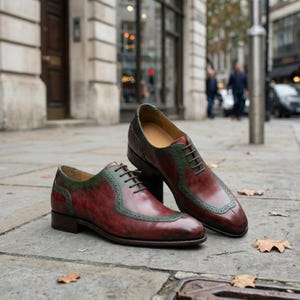 May include: A pair of men's leather dress shoes in burgundy and green. The shoes feature a classic Oxford design with a cap toe and decorative stitching. The shoes are displayed on a pavement in a city setting.