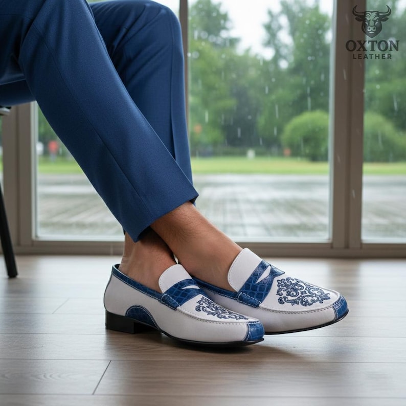 May include: White and blue leather loafers with intricate floral embroidery. The shoes feature a white base with blue accents on the trim and a decorative strap. The design includes a black sole and a classic loafer silhouette. The shoes are from Oxton Leather.